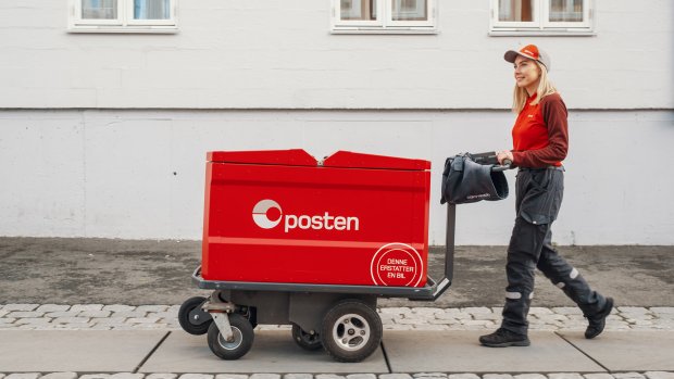 Kvinnelig postbud går i bygata med elektrisk posttralle