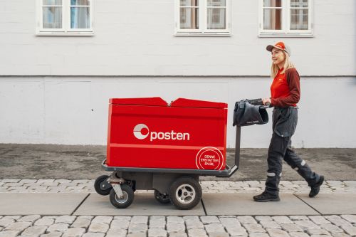 Kvinnelig postbud går i bygata med elektrisk posttralle