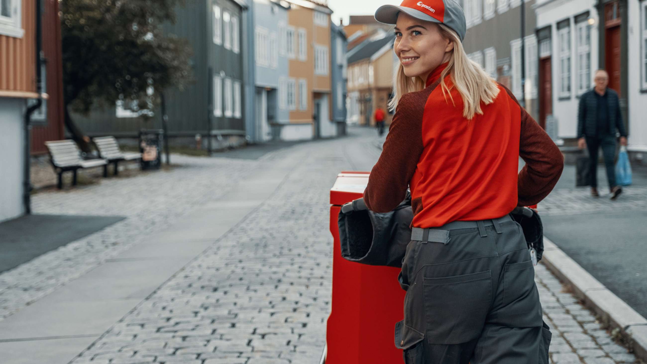 Postbud smiler over ryggen med tralle i gatemiljø